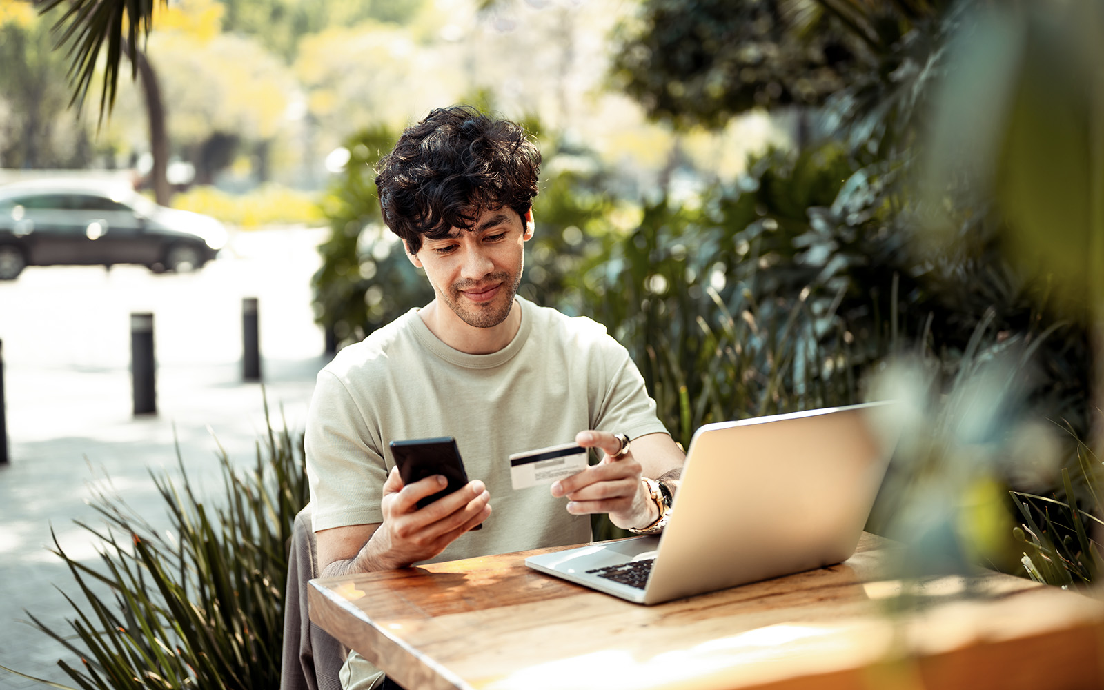 A man sitting at an outdoor table with a laptop, looking at his phone and credit card, as if about to use some form of online marketplace