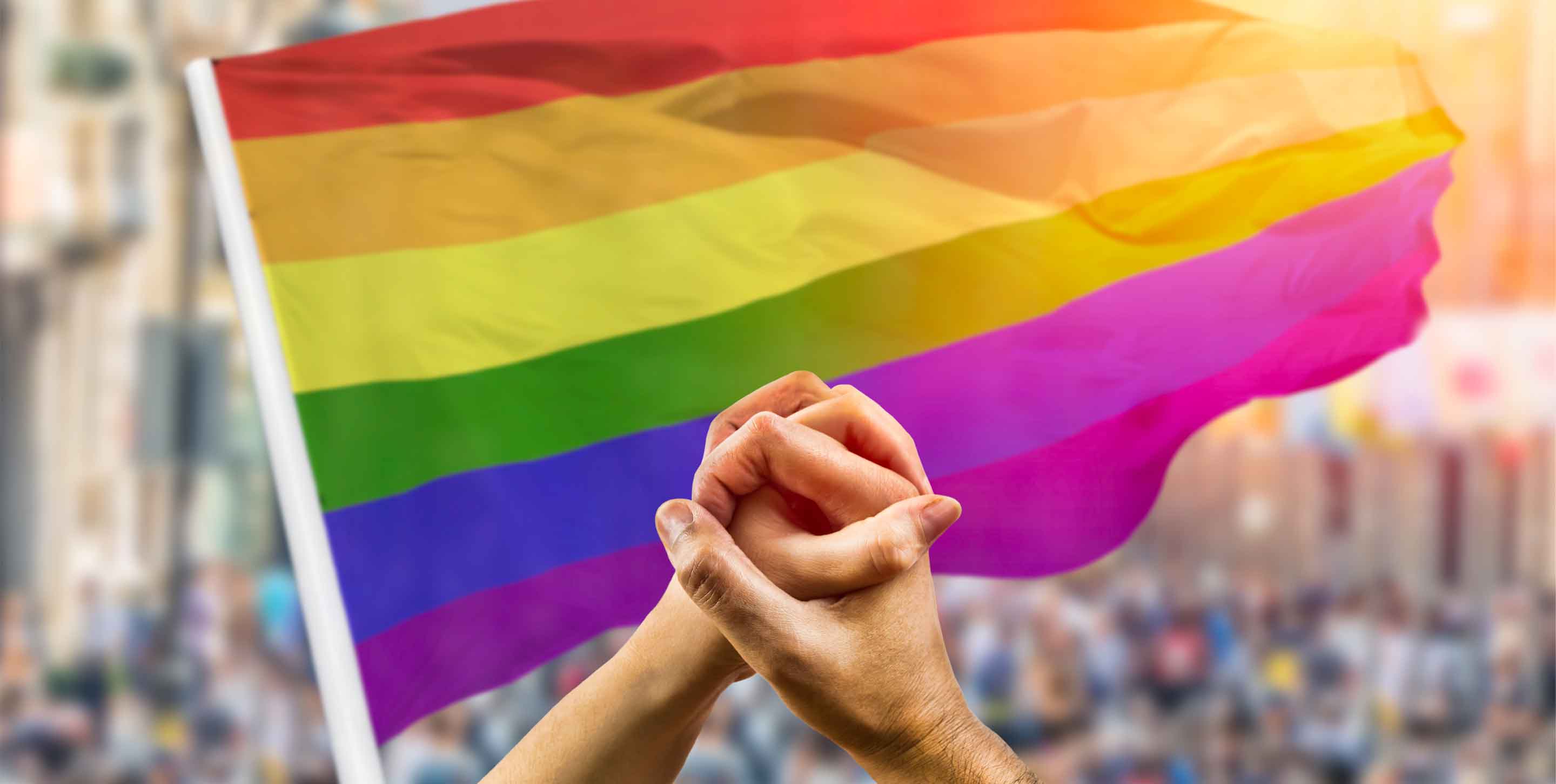 Cropped shot of a couple holding hands and wave in front of a rainbow flag flying on the sidelines of a summer gay pride parade