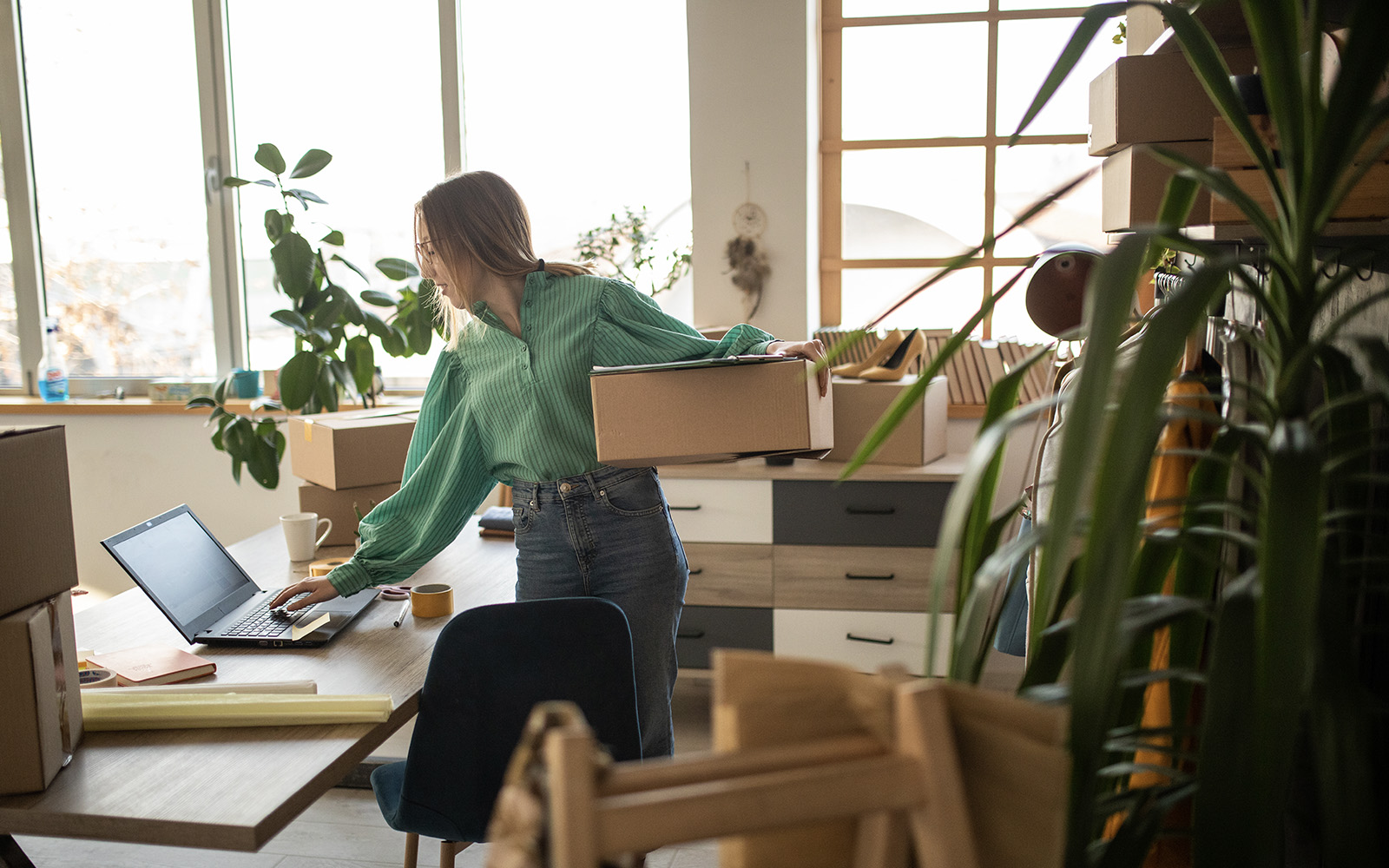 A far-shot of a woman standing at her desk, one hand on her laptop to take care of her ecommerce site. Her other arm holds a box, as if preparing to ship an order. Her office is filled with similar boxes, books, and greenery