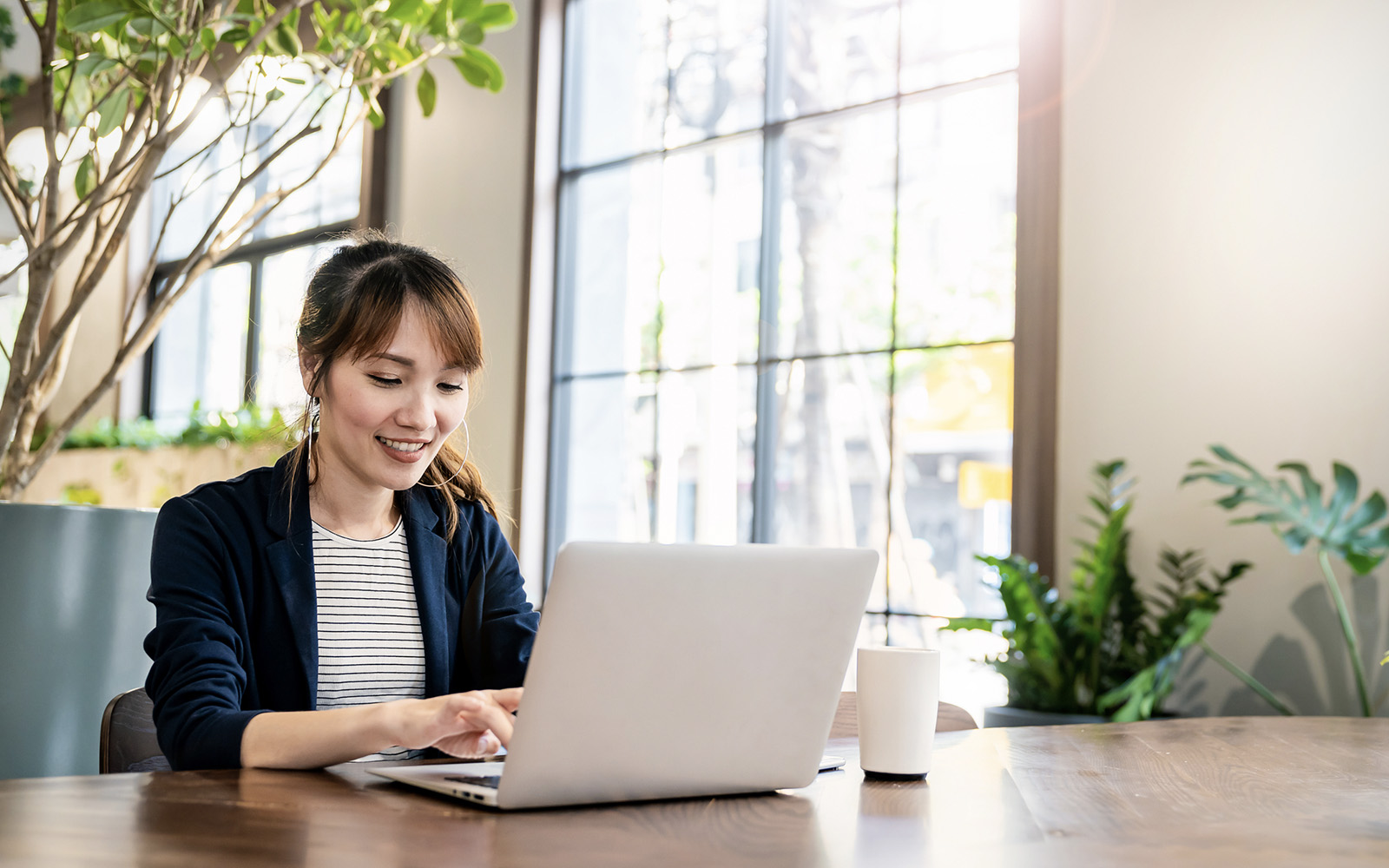 A woman using Vertex Certificate Center on her laptop. She sits at a desk, the room brightly lit by large windows in the background, and several plants appear interspersed throughout the room