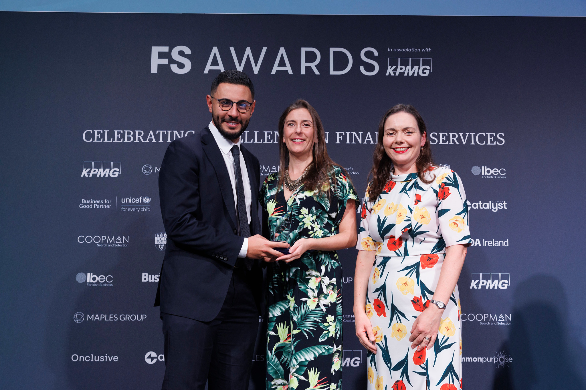 Image of three people in front of a blue background that reads "FS AWARDS. In association with KPMG. Celebrating excellence in financial services." One person in a suit is presenting the award to the other two, who wear brightly-colored dresses. All three are smiling.