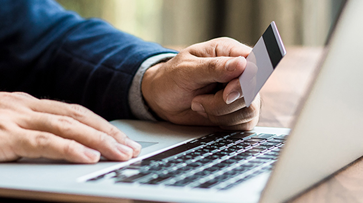 A close-up image of hands resting on the keyboard of an open laptop. One hand holds a credit card to examine the numbers on the back. The person who the hands belong to appears to be wearing a blue suit.