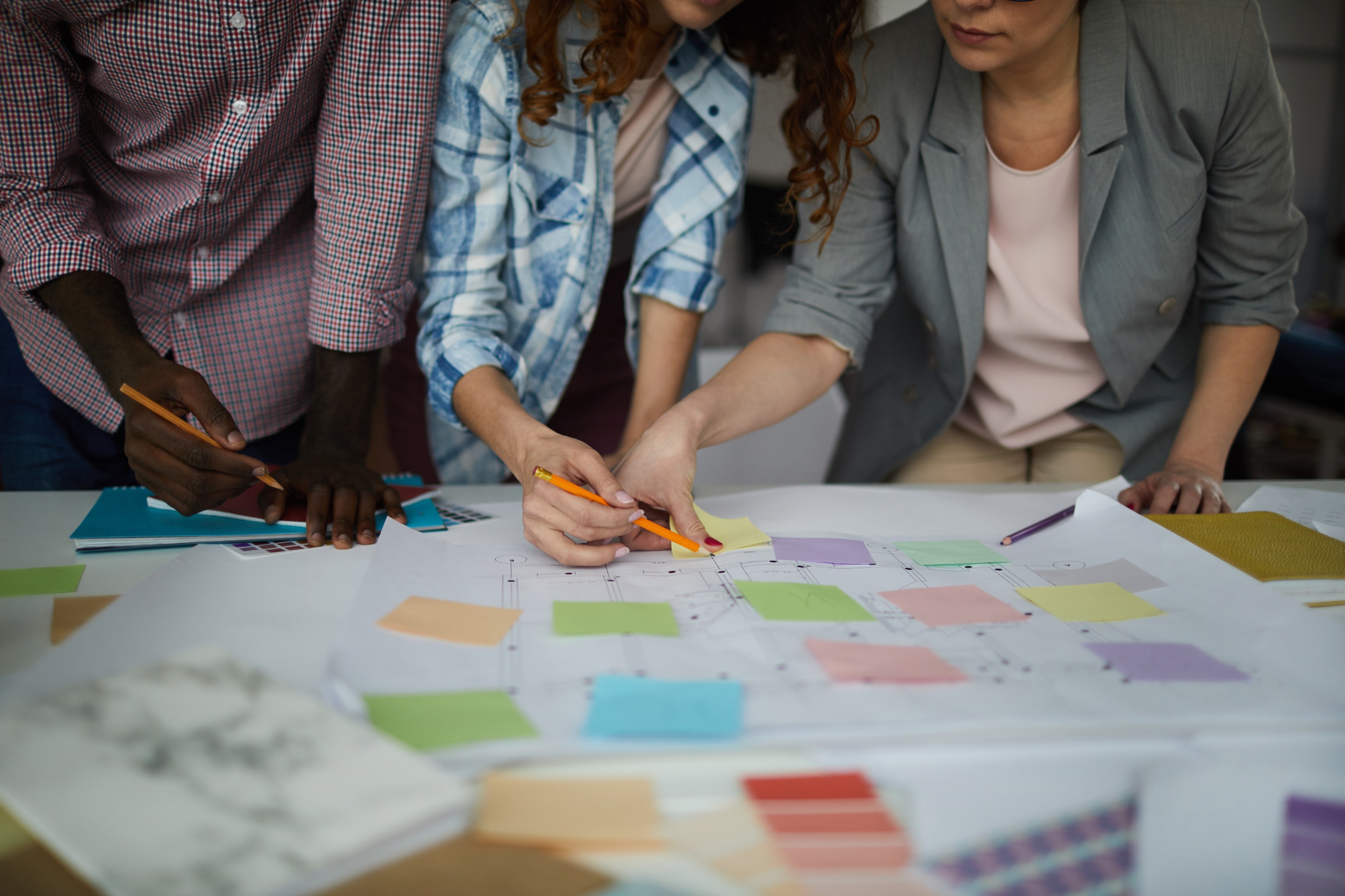 A team of collaborators leans over plans laid out across a desk in front of them. The desk is filled with notebooks, planning documents, and colorful sticky notes that are placed to help organize the work. Three people, shown from the neck down, collaborate on a work with pencils in hand to show they are taking an active role in their planning process.