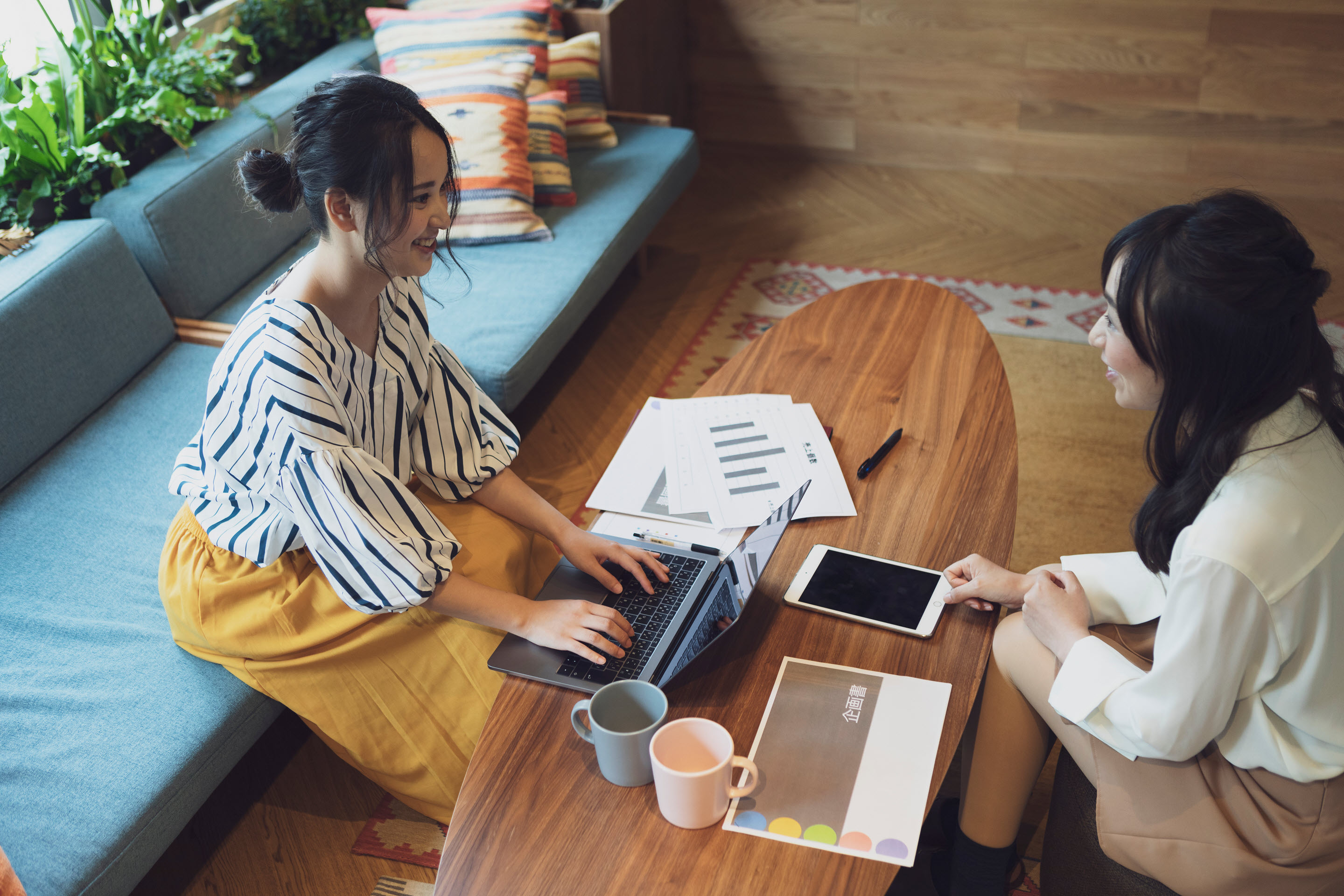 Two businesswomen in casual dress sit across from one another in a cozy lounge area. They both have laptops, phones, and paperwork on the coffee table as they work.