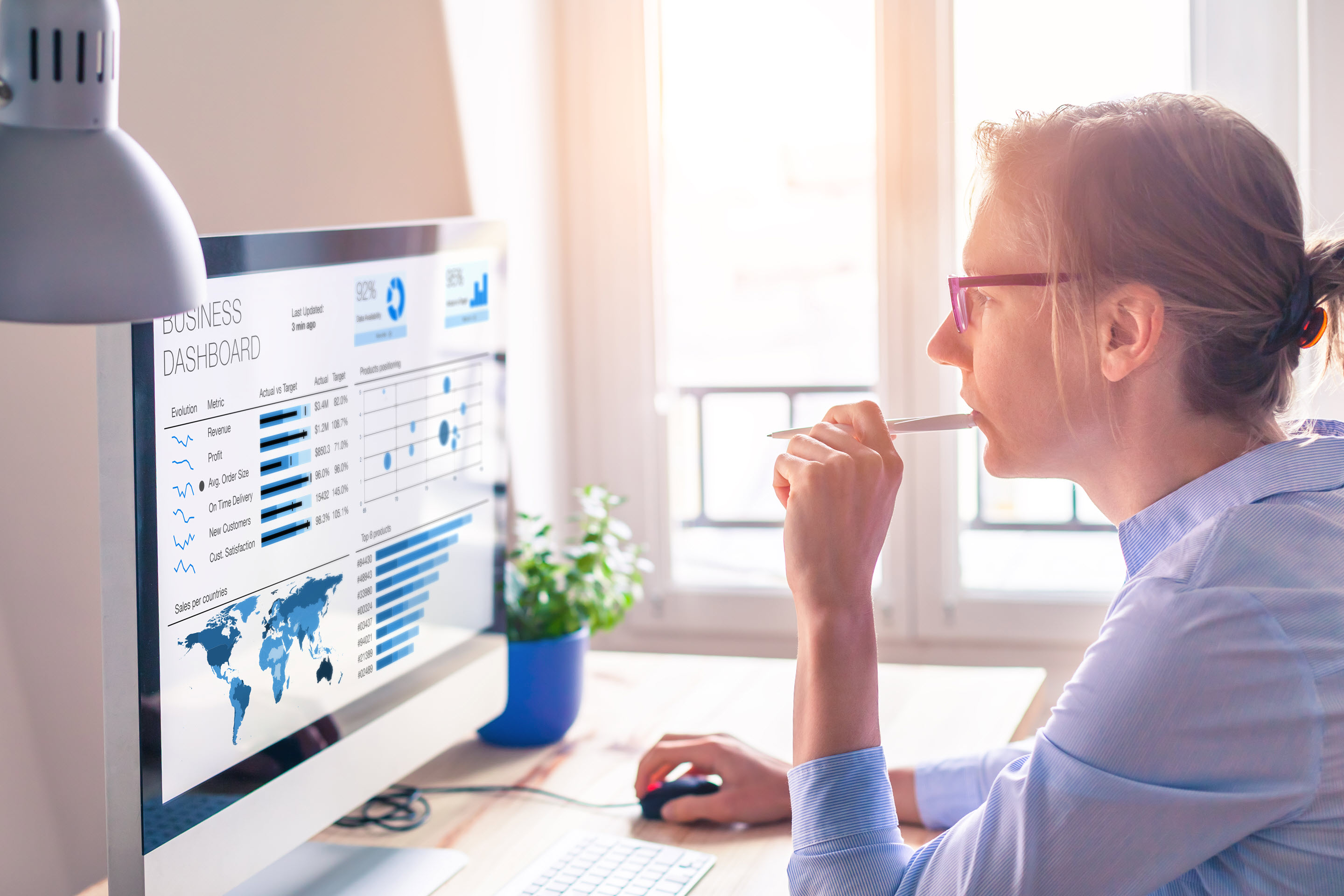 A woman in thought at her computer. She is reviewing a global business dashboard on her computer in front of her with various data metrics and blue graphs related to her organization. She is chewing on a pen, has her glasses on, and has her hair in a bun to illustrate her attentiveness.