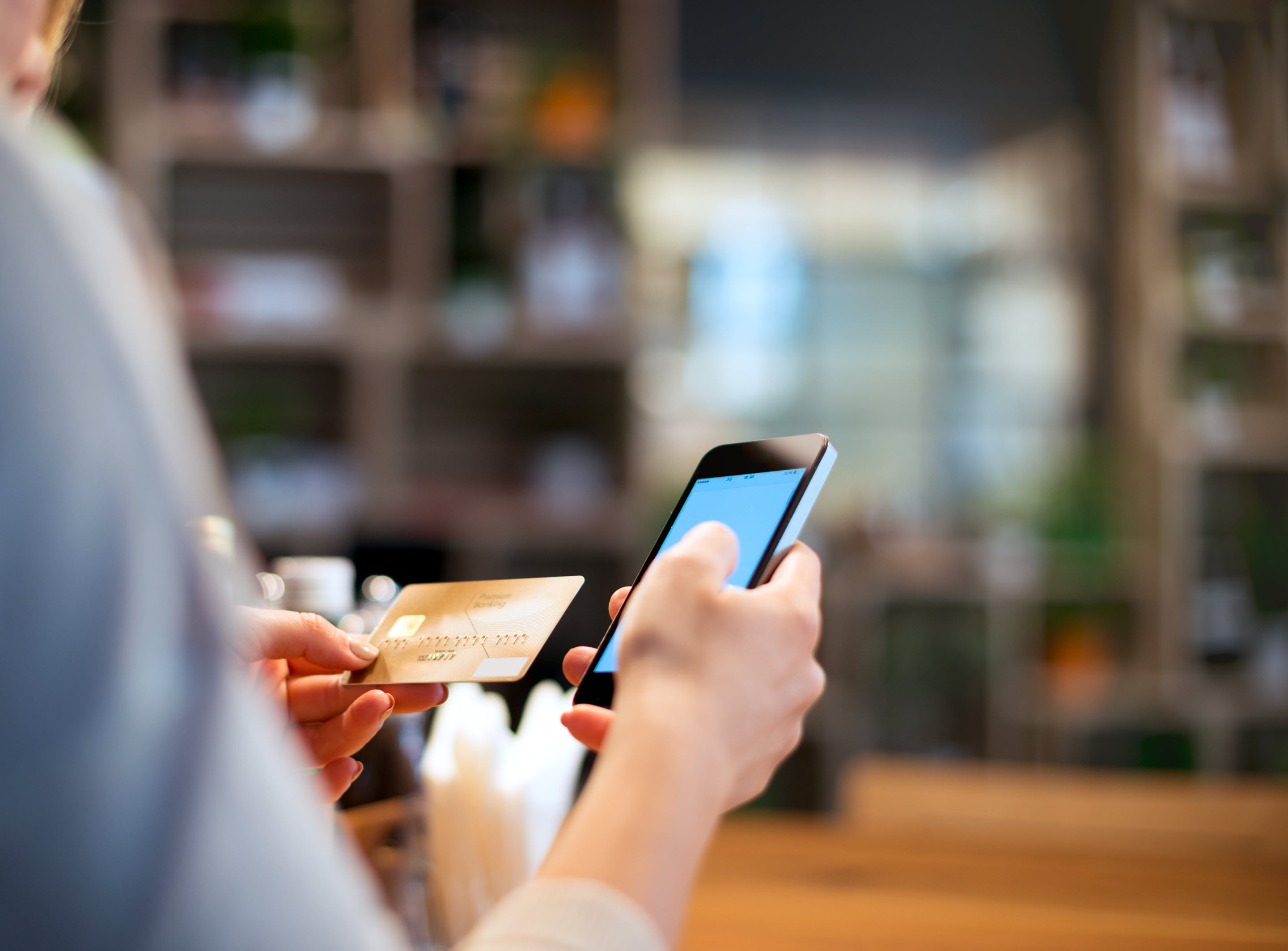 Over-the-shoulder shot of someone examining their credit card in one hand with their phone in the other. The background is comprised of blurred shelves that suggest a shop environment.