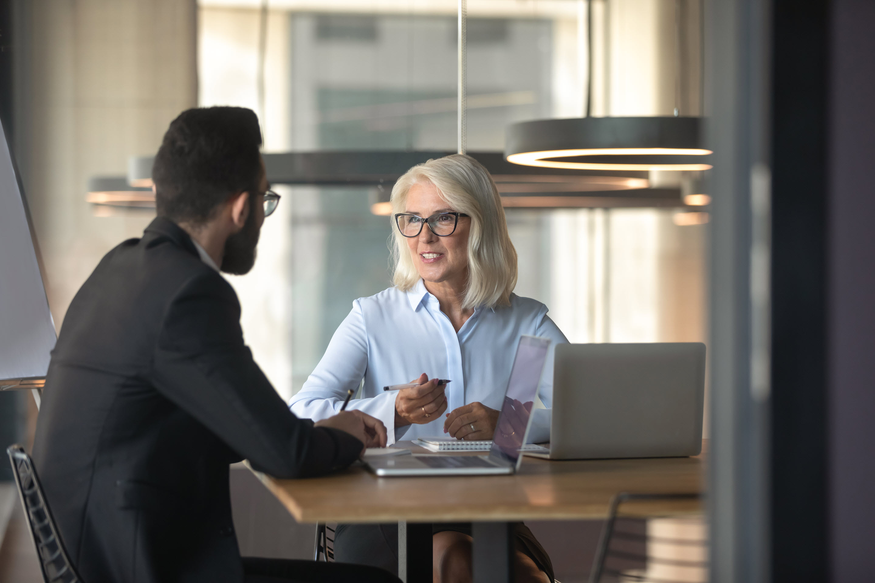 A woman and a man in business attire sit at a table in a well-lit room, discussing data and tax compliance insights from the laptop that rests in front of the woman.