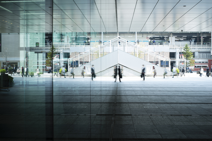 A pedestrian walkway, as seen from the underside of a glass walkway. Several business people walk about the scene with purpose.