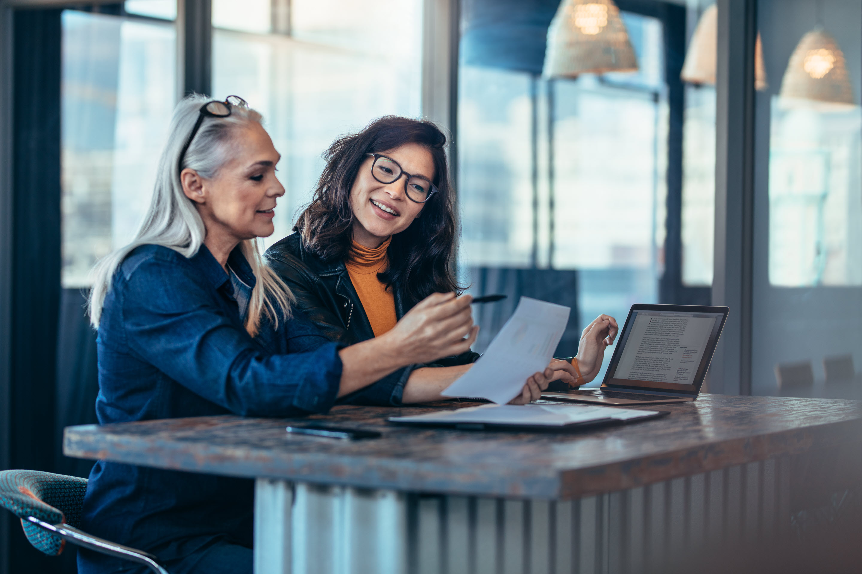 Two businesswomen sit next to each other in a well-lit office space. They both have laptops and are discussing something based on the data on their devices..