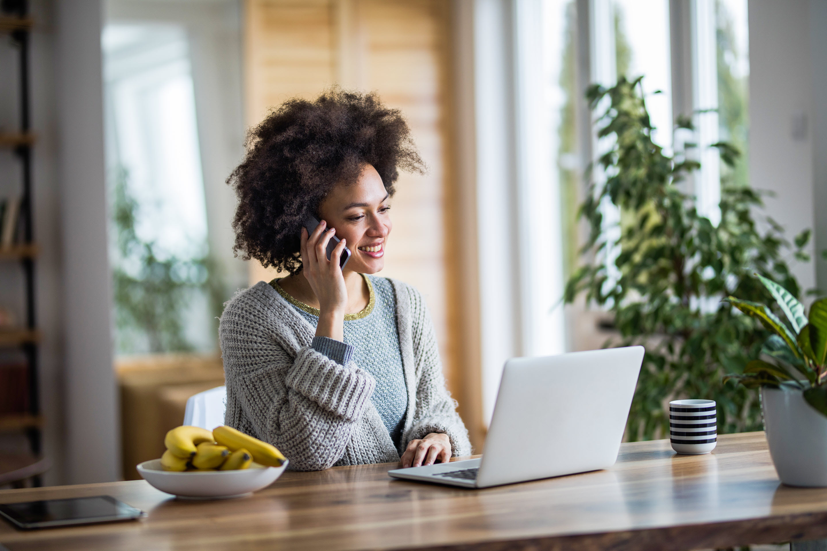 A woman browses Vertex solutions on her laptop while talking on the phone, in the comfort of her home. She is surrounded by greenery.