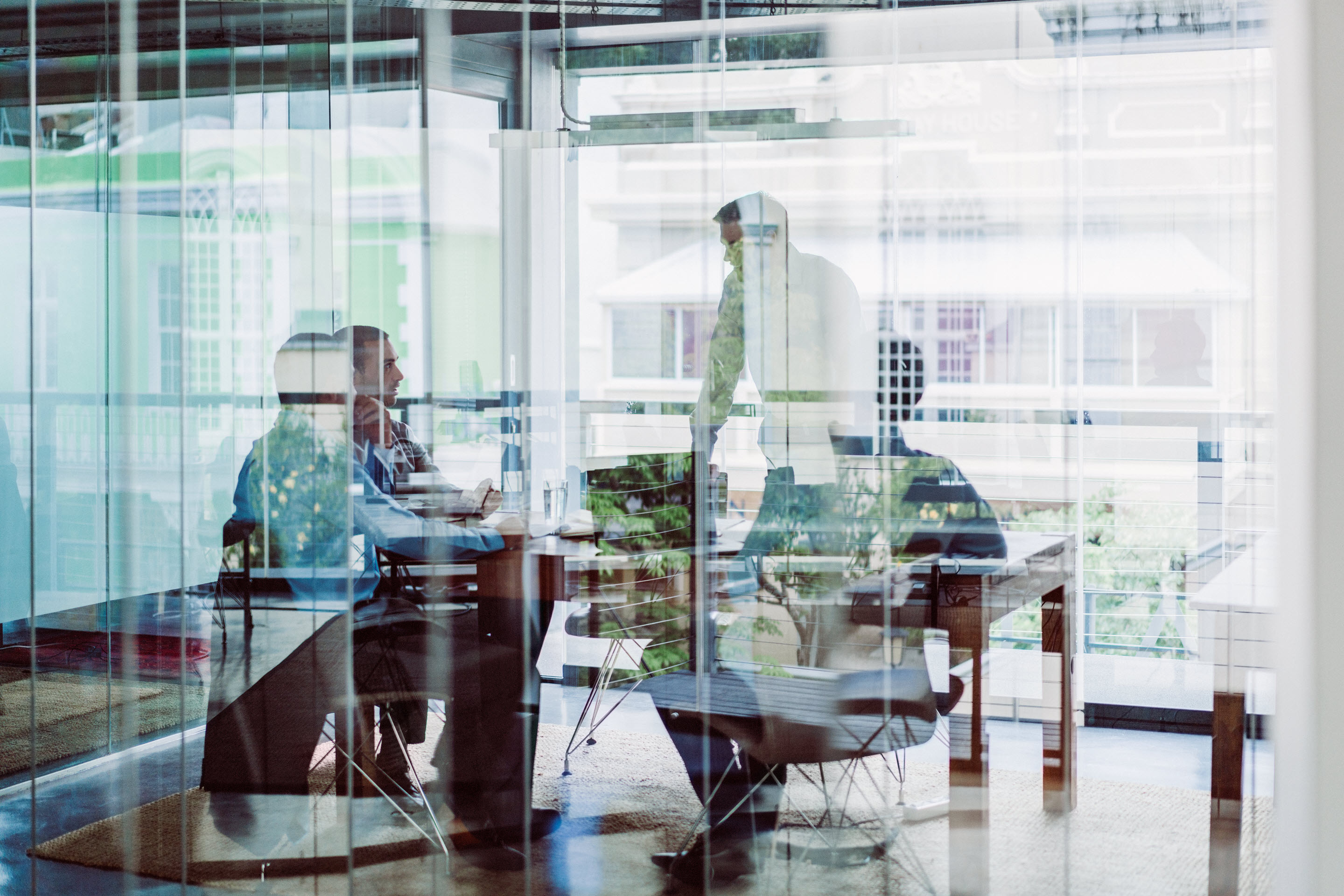 A group of business executives and thought leaders gather around a conference table for discussion. Part of the image is overlaid and obscured by the blue tone of reflection from the glass wall of the room.