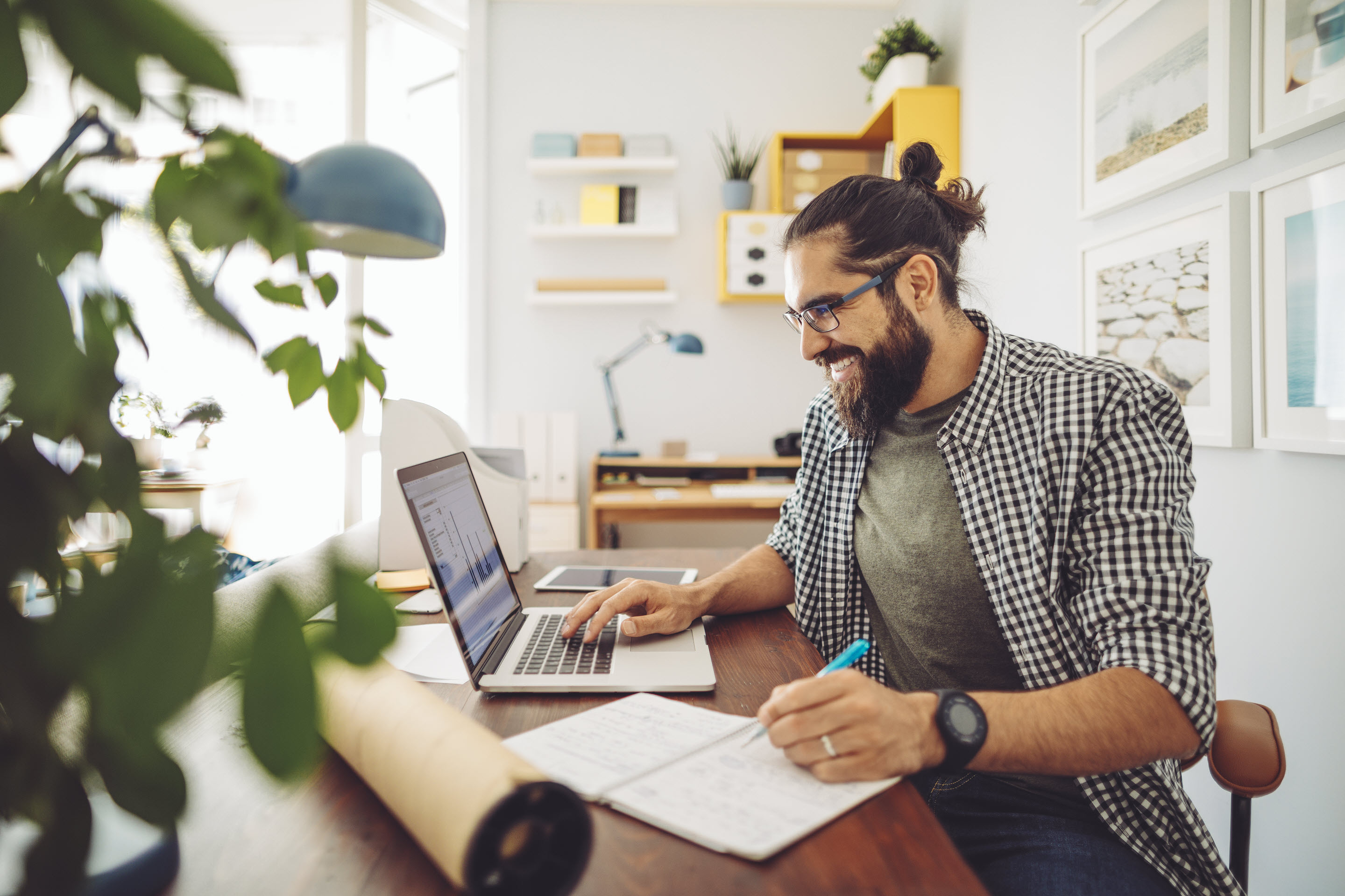 A man works from home, taking notes in front of his laptop. The office is entirely comprised of light colors, making it feel peaceful. A plant is visible but blurred in the foreground.