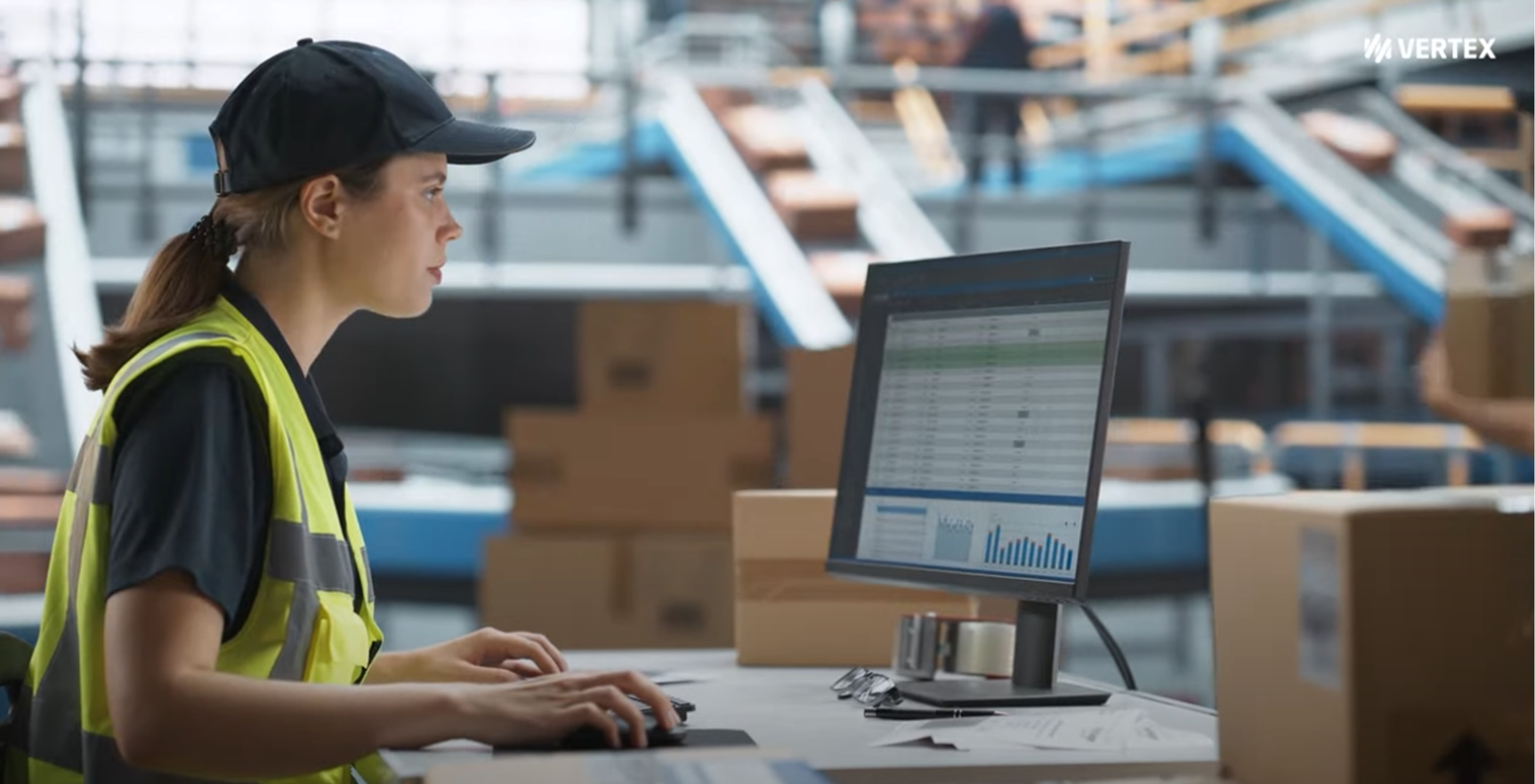 A woman wearing a high-vis vest sits in front of a computer, the desk full of boxes. In the background, there is an out-of-focus conveyer belt with more of the boxes that are on the table.