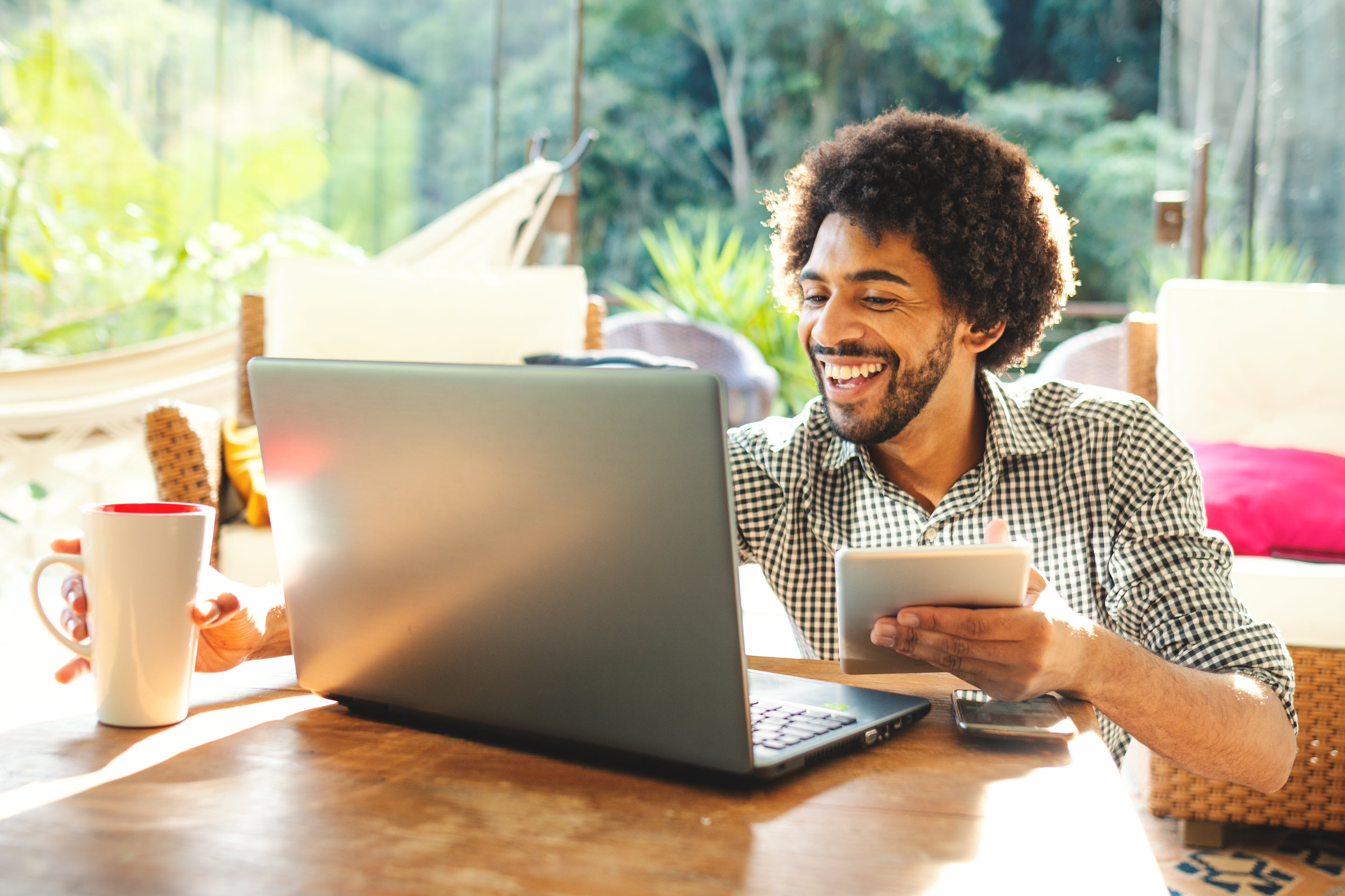 A smiling man sits at his computer, with a large open window showing vibrant greenery behind him.