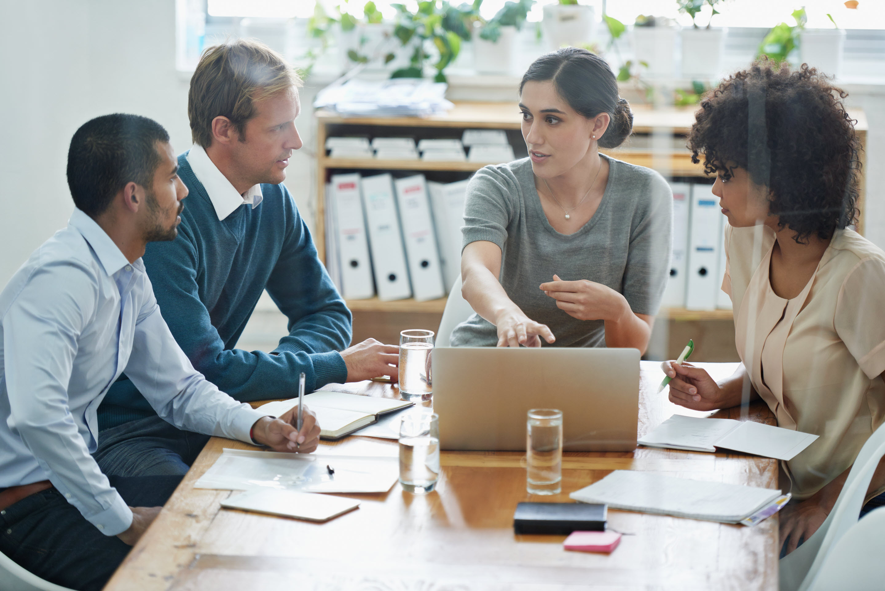 A businesswoman explains issues to her staff at their collaborative work desk, surrounded by files and greenery.