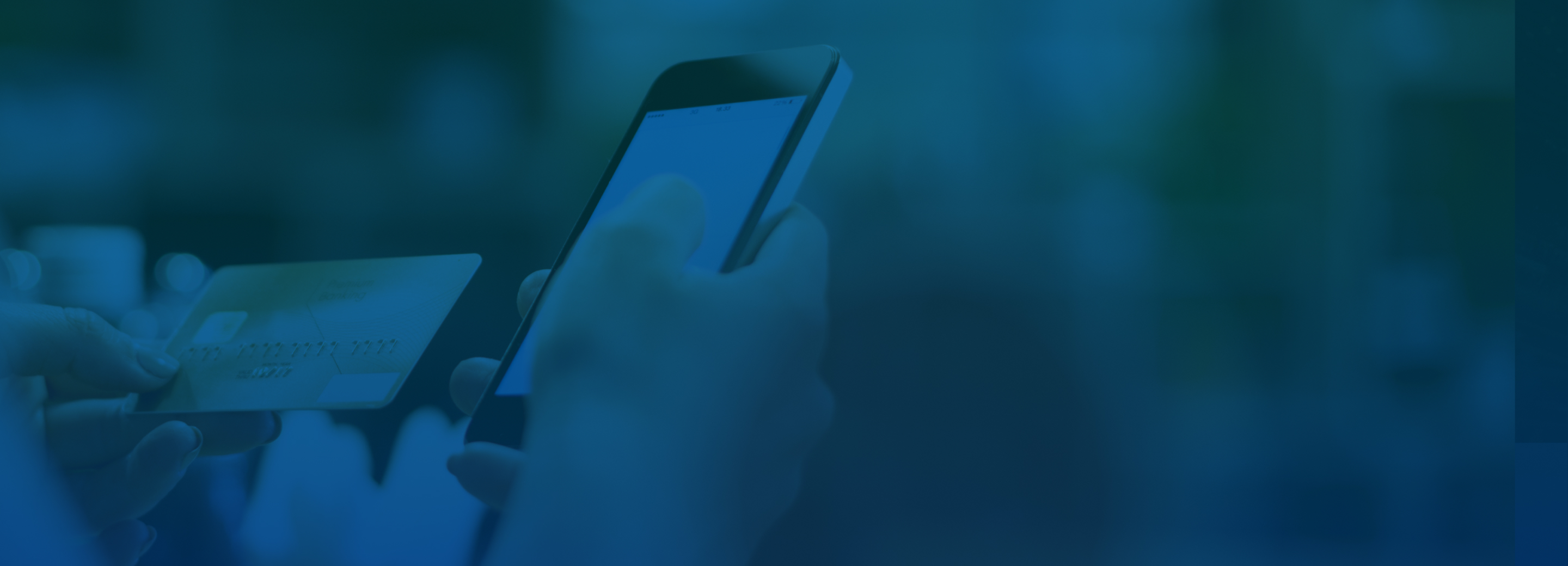 Over-the-shoulder shot of someone examining their credit card in one hand with their phone in the other. The background is comprised of blurred shelves that suggest a shop environment. A blue overlay has been added to the image.