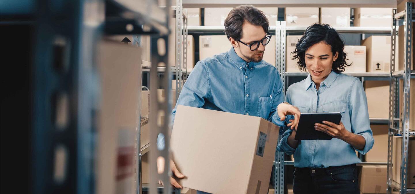 Female Inventory Manager Shows Digital Tablet Information to a Worker Holding Cardboard Box, They Talk and Do Work. In the Background Stock of Parcels with Products Ready for Shipment.