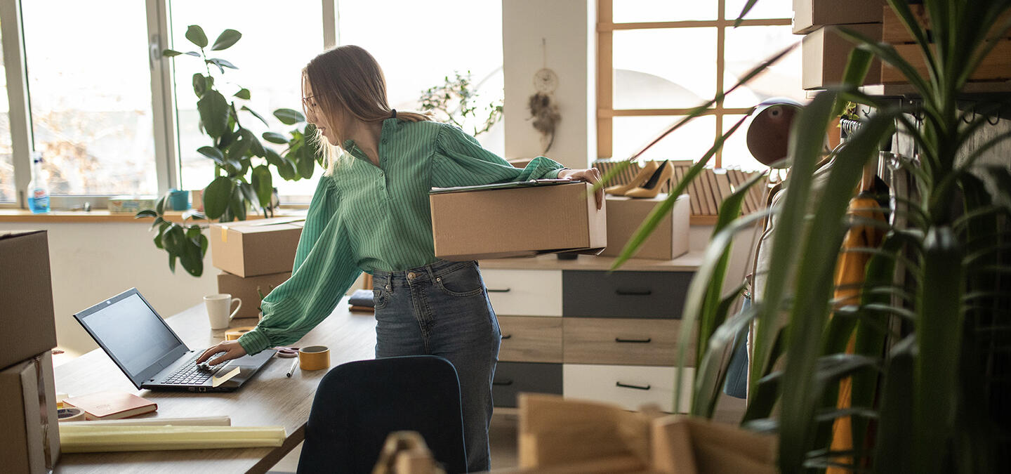 A far-shot of a woman standing at her desk, one hand on her laptop to take care of her ecommerce site. Her other arm holds a box, as if preparing to ship an order. Her office is filled with similar boxes, books, and greenery
