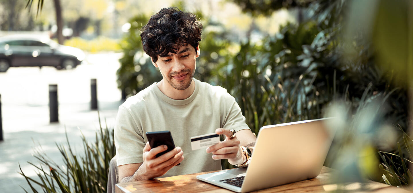 A man sitting at an outdoor table with a laptop, looking at his phone and credit card, as if about to use some form of online marketplace