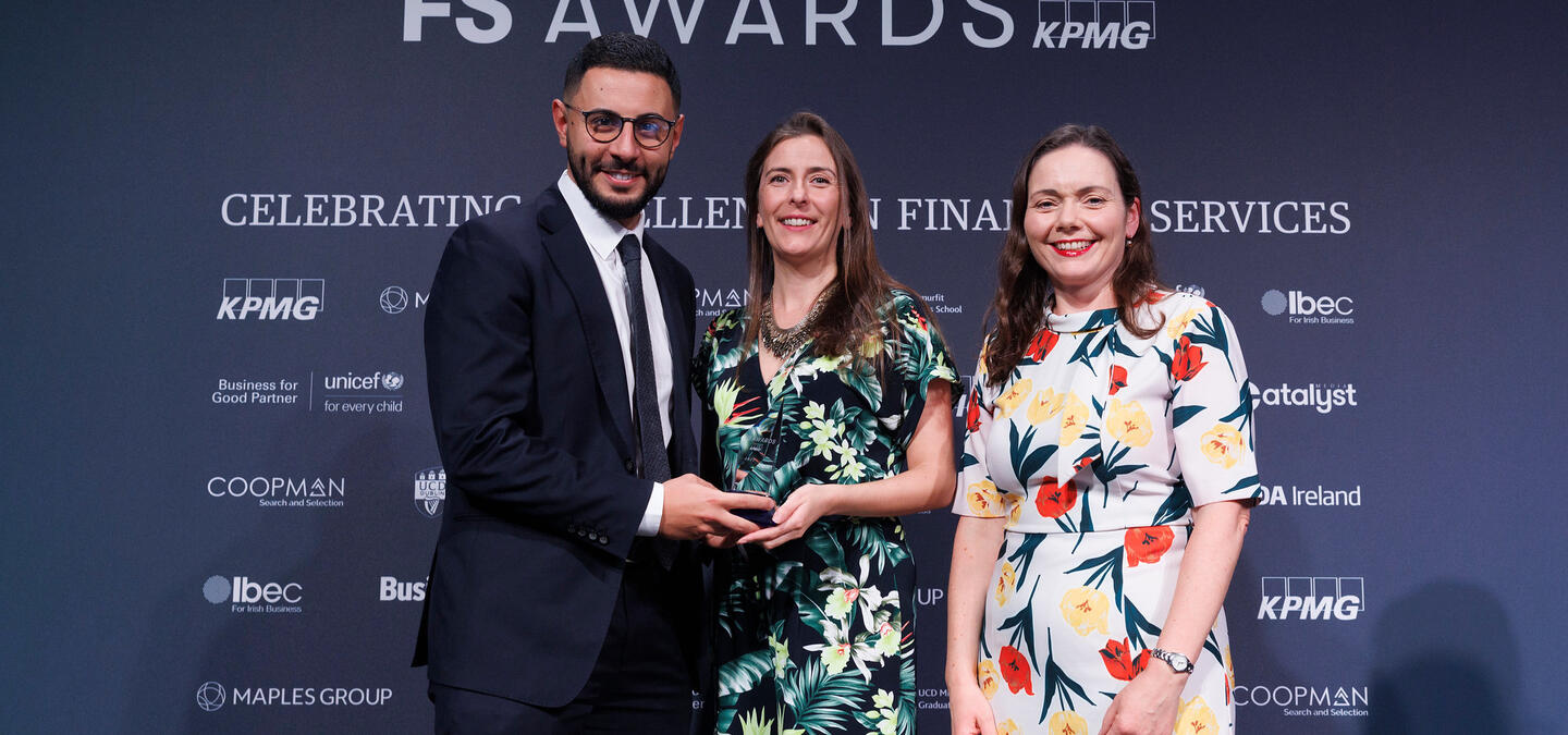 Image of three people in front of a blue background that reads "FS AWARDS. In association with KPMG. Celebrating excellence in financial services." One person in a suit is presenting the award to the other two, who wear brightly-colored dresses. All three are smiling.
