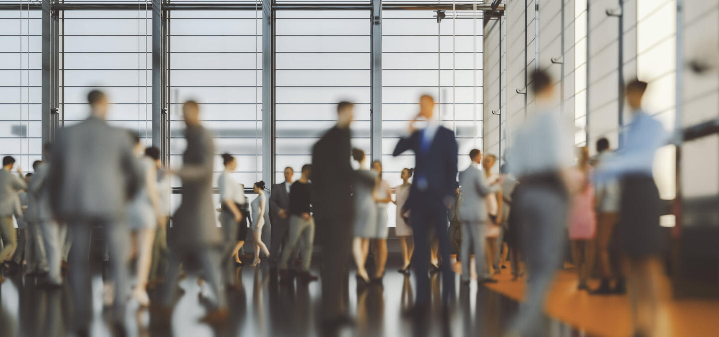 A conference room with large floor-to-ceiling windows, allowing natural light in. A crowd of businesspeople in formal clothing takes up most of the image, with many of the people interacting with one another: holding conversation, shaking hands, et cetera. The people are all blurred, as if they are in motion.