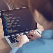 An over-the-shoulder image of a woman sat at a warm wooden table, looking over code on a laptop.