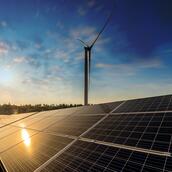 Solar panels approach the camera from a diagonal, reflecting bright yellow light. Above is a deep blue sky with a wind turbine.