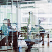 A group of business executives and thought leaders gather around a conference table for discussion. Part of the image is overlaid and obscured by the blue tone of reflection from the glass wall of the room.