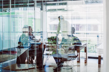 A group of business executives and thought leaders gather around a conference table for discussion. Part of the image is overlaid and obscured by the blue tone of reflection from the glass wall of the room.