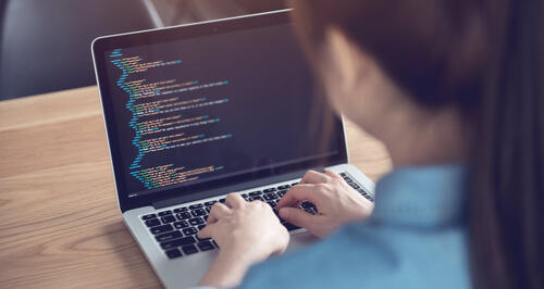 An over-the-shoulder image of a woman sat at a warm wooden table, looking over code on a laptop.