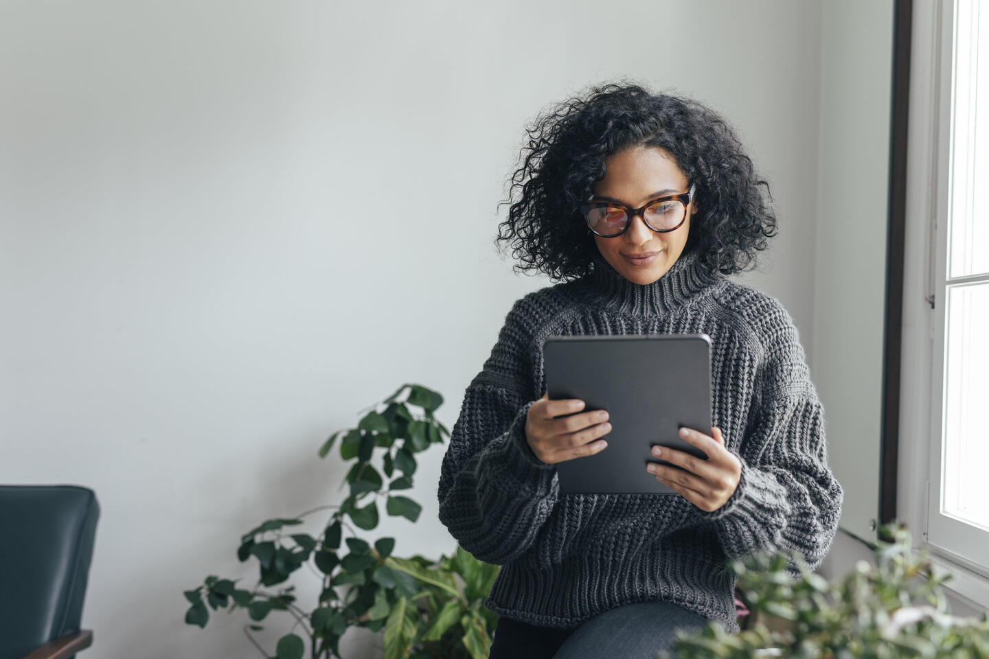 A woman stands by a window, surrounded by plants. She looks down at her tablet to monitor her e-invoices, audits, and reports from Vertex e-Invoicing.