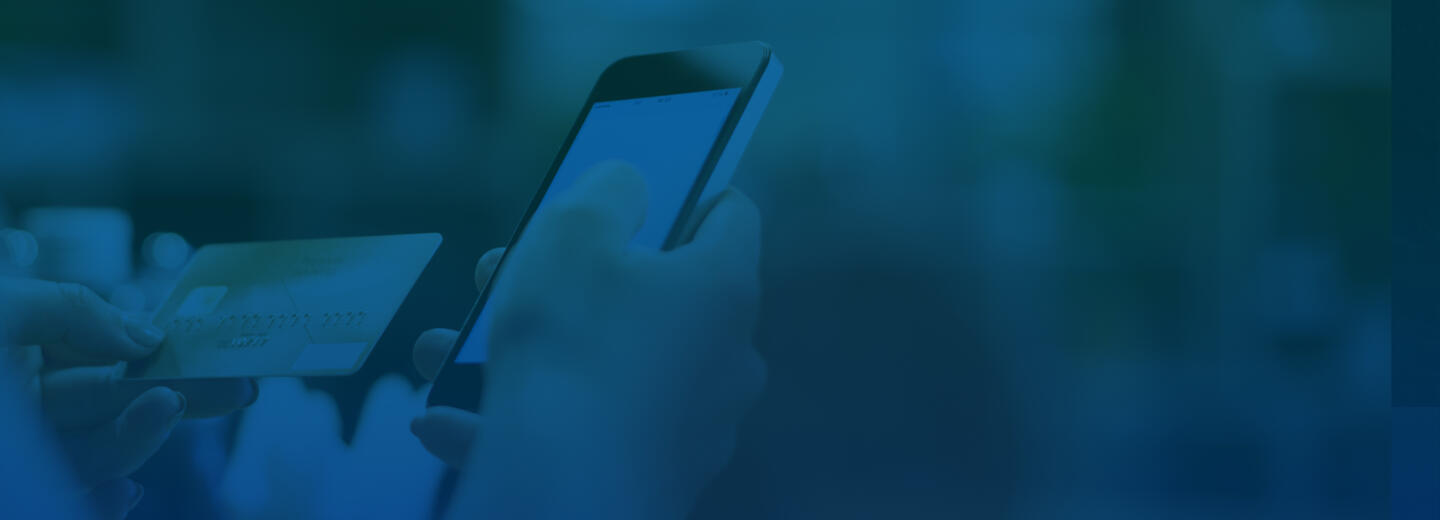 Over-the-shoulder shot of someone examining their credit card in one hand with their phone in the other. The background is comprised of blurred shelves that suggest a shop environment. A blue overlay has been added to the image.