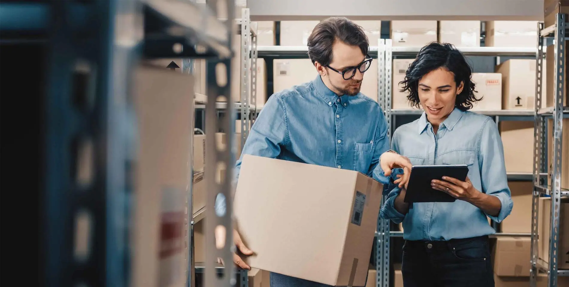 Female Inventory Manager Shows Digital Tablet Information to a Worker Holding Cardboard Box, They Talk and Do Work. In the Background Stock of Parcels with Products Ready for Shipment.