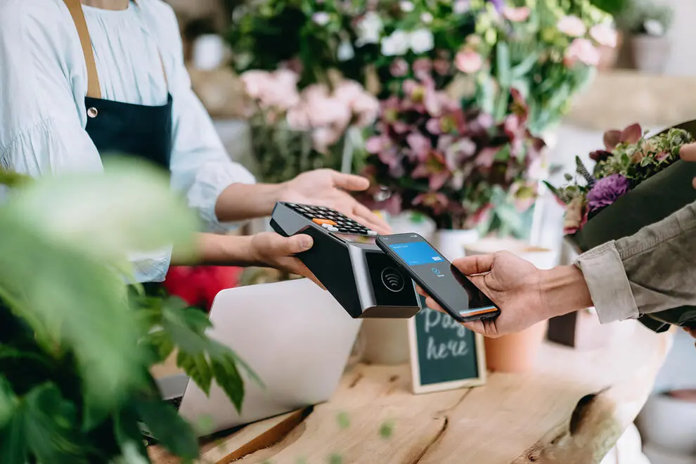 Two hands performing an e-commerce transaction using the customer's phone. Flowers are visible around the shop and in the customer's arms, which is presumably what they are purchasing. The shopkeeper is partially obscured by a green plant in the foreground