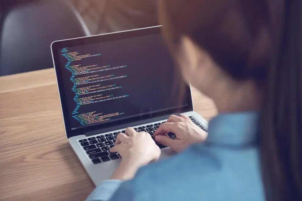 An over-the-shoulder image of a woman sat at a warm wooden table, looking over code on a laptop.