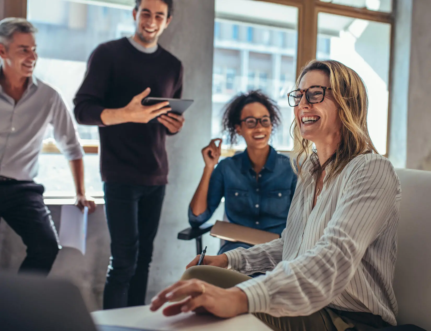 Four people in business-casual attire are an office space, which has big sunny windows. The woman seated closest to the camera is referencing and reaching for a computer in front of her. She is smiling with the three people, slightly out of focus behind her, who are all taking a look at the work in front of her.