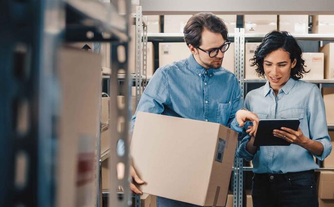 Female Inventory Manager Shows Digital Tablet Information to a Worker Holding Cardboard Box, They Talk and Do Work. In the Background Stock of Parcels with Products Ready for Shipment.