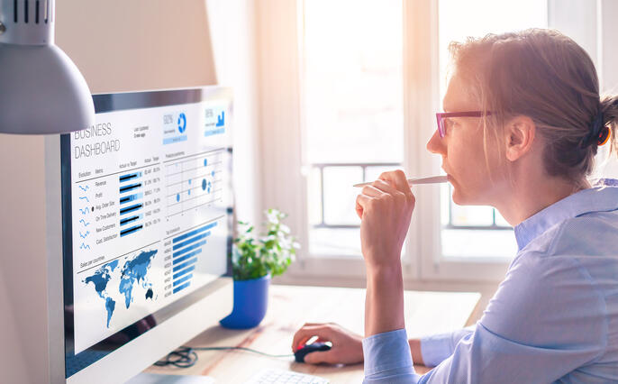 A woman in thought at her computer. She is reviewing a global business dashboard on her computer in front of her with various data metrics and blue graphs related to her organization. She is chewing on a pen, has her glasses on, and has her hair in a bun to illustrate her attentiveness.