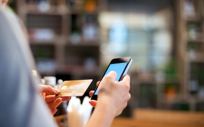 Over-the-shoulder shot of someone examining their credit card in one hand with their phone in the other. The background is comprised of blurred shelves that suggest a shop environment.