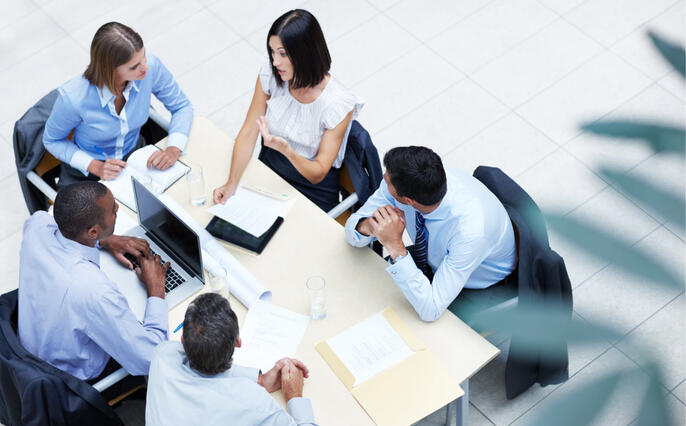 A group of business professionals with laptops, leaning in to a table mid-conversation. The table is shot from a birds-eye view, with a plant blurred in the periphery of the bottom right corner of the image.