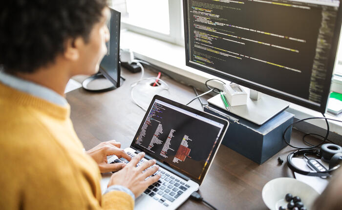 An over-the-shoulder view of a senior IT manager, dressed in yellow. They are working at their laptop, plugged into a monitor behind it, and looking at raw data for tax insights.