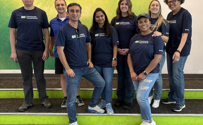 Eight Vertex employees, wearing matching Community Connections shirts, volunteer. They stand on three small steps, with a large mural of a green apple in the background.