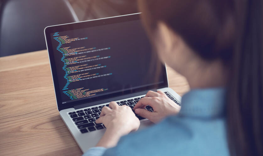 An over-the-shoulder image of a woman sat at a warm wooden table, looking over code on a laptop.