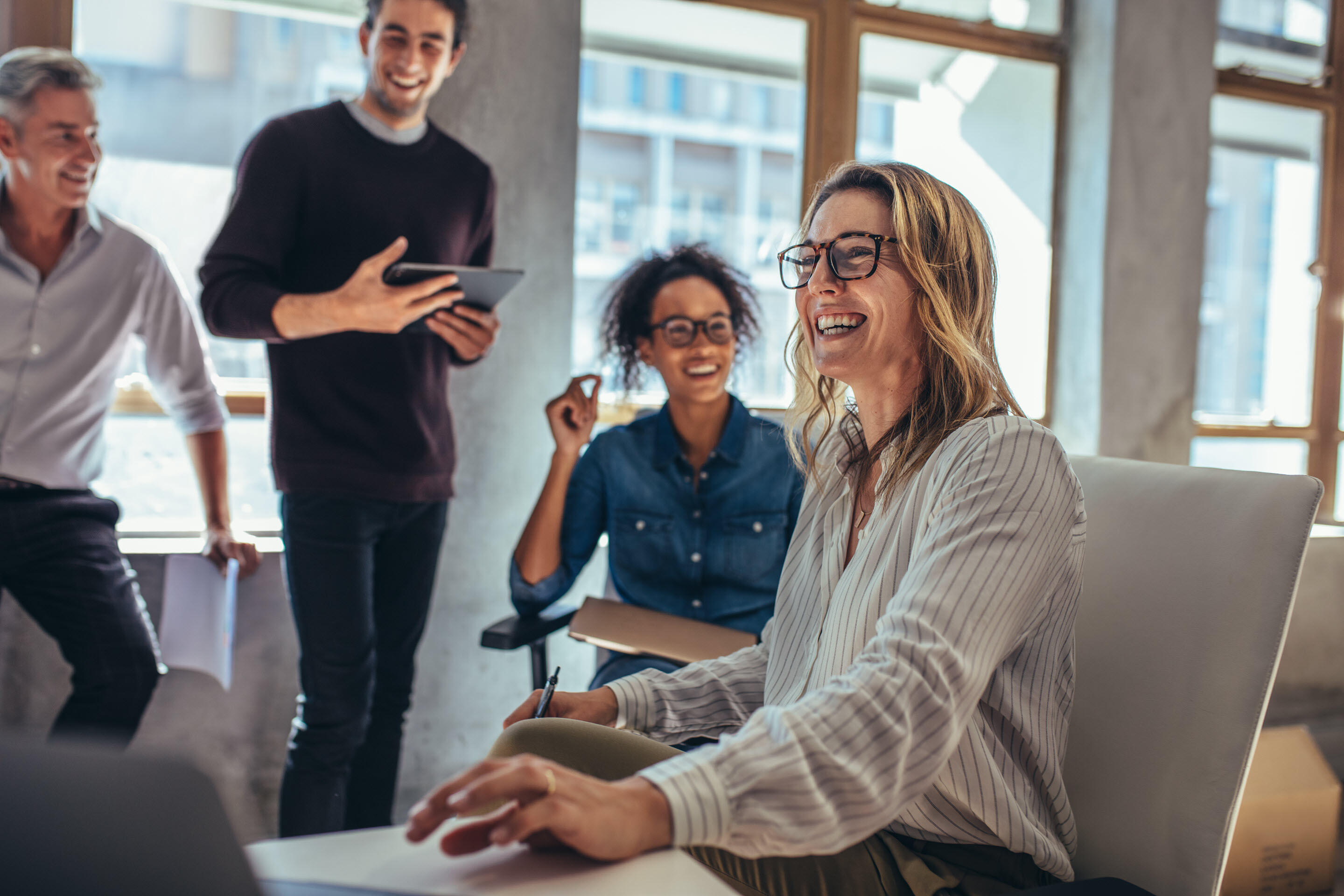 Four people in business-casual attire are an office space, which has big sunny windows. The woman seated closest to the camera is referencing and reaching for a computer in front of her. She is smiling with the three people, slightly out of focus behind her, who are all taking a look at the work in front of her.