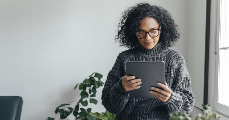 A woman stands by a window, surrounded by plants. She looks down at her tablet to monitor her e-invoices, audits, and reports from Vertex e-Invoicing.